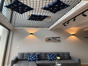 Una sala de estar moderna con un sofá gris, cojines estampados, una mesa de centro de vidrio con un jarrón y un cuadro con temática náutica en la pared.