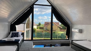 Un pequeño dormitorio en el ático, con techo inclinado, un ventanal y vistas a casas y vegetación.