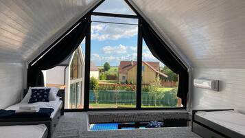 A room with a sloped ceiling, a bed with a mattress and pillows, a window with a view of houses and greenery, and a small pool.