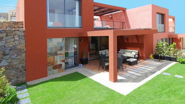 Ein modernes, zweistöckiges Haus mit Steinmauer, einer Terrasse mit Gartenmöbeln und einem gepflegten Rasen.