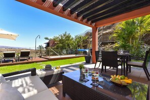 Une terrasse extérieure avec une table en verre, des meubles en osier et une vue sur une piscine et un espace verdoyant.