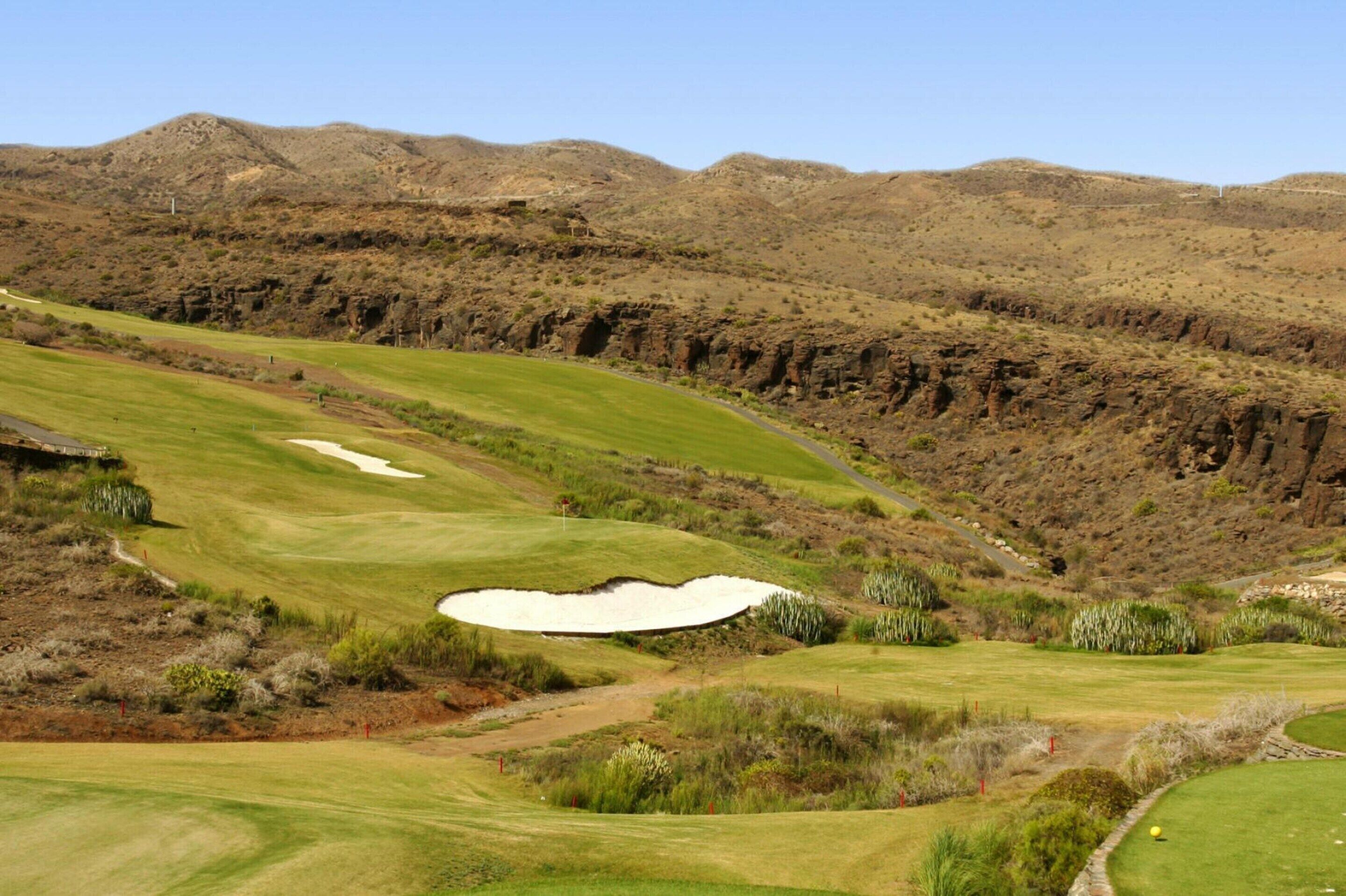 A golf course with bunkers, a water hazard, and a mountainous backdrop.