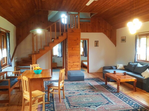 Ein gemütliches Blockhaus-Interieur mit einer Holztreppe, einem Essbereich mit Tisch und Stühlen, einer Couch und einem gemusterten Teppich.