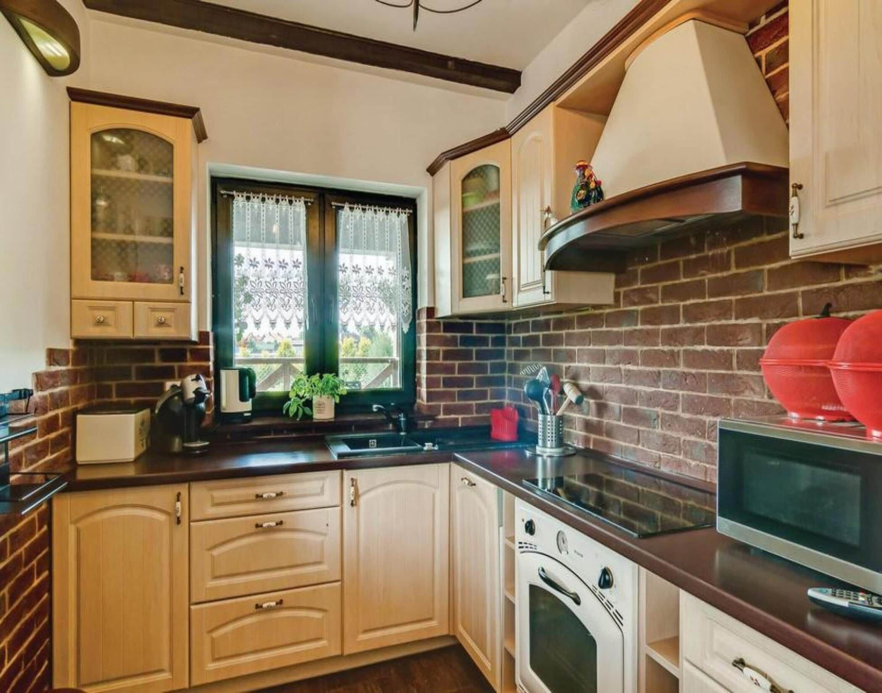 A kitchen with a brick wall, wooden cabinets, a window with lace curtains, and a microwave.