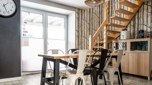 Un comedor con mesa y sillas de madera, un reloj en la pared y una escalera con papel tapiz de motivos arbóreos.