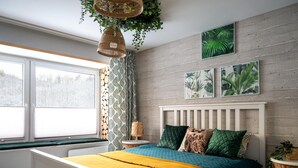 Un dormitorio con una cama blanca, una manta amarilla, ropa de cama verde y azul, un cabecero de madera y una ventana con vistas a árboles.