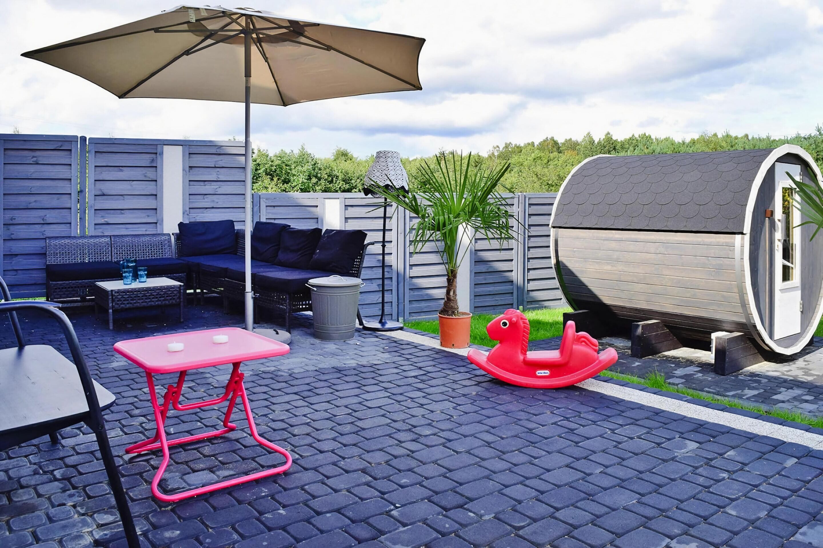 An outdoor patio area with a sofa set, a table, an umbrella, a planter, a rocking horse, and a cylindrical structure.