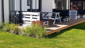 Ein moderner Außenbereich mit Holzterrasse, weißen Möbeln und einer Glastür mit der Aufschrift „7.2“.