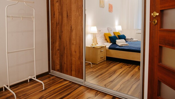 A hotel room with a wooden floor, a bed with blue and yellow pillows, a nightstand with a lamp, and a wooden wardrobe. - Apartment in Lukecin Near Baltic Sea (Dziwnów)
