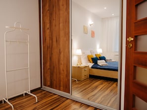 A hotel room with a wooden floor, a bed with blue and yellow pillows, a nightstand with a lamp, and a wooden wardrobe. - Apartment Close to the Beach, Summer Pool (Dziwnów)