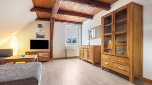 Ein gemütliches Wohnzimmer mit Holzbalkendecke, einem Flachbildfernseher, einer Couch, einem Esstisch und einer Vitrine mit Glastüren.
