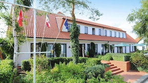 A two-story building with a red-tiled roof, white walls, and multiple windows. There are flags hanging from a flagpole, and a green umbrella is visible in the foreground. - Appartements Post Hiddensee in Vitte (Hiddensee)