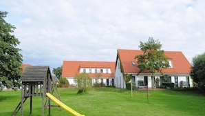 A playground with a yellow slide and a wooden playhouse, set against a backdrop of two white houses with red-tiled roofs. - Appartements Post Hiddensee in Vitte (Hiddensee)