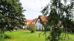A white building with a red roof, surrounded by a well-maintained lawn and trees. - Appartements Post Hiddensee in Vitte (Hiddensee)