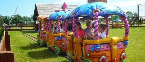 A colorful train ride with passengers, set on a grassy track with a wooden fence and a building in the background.