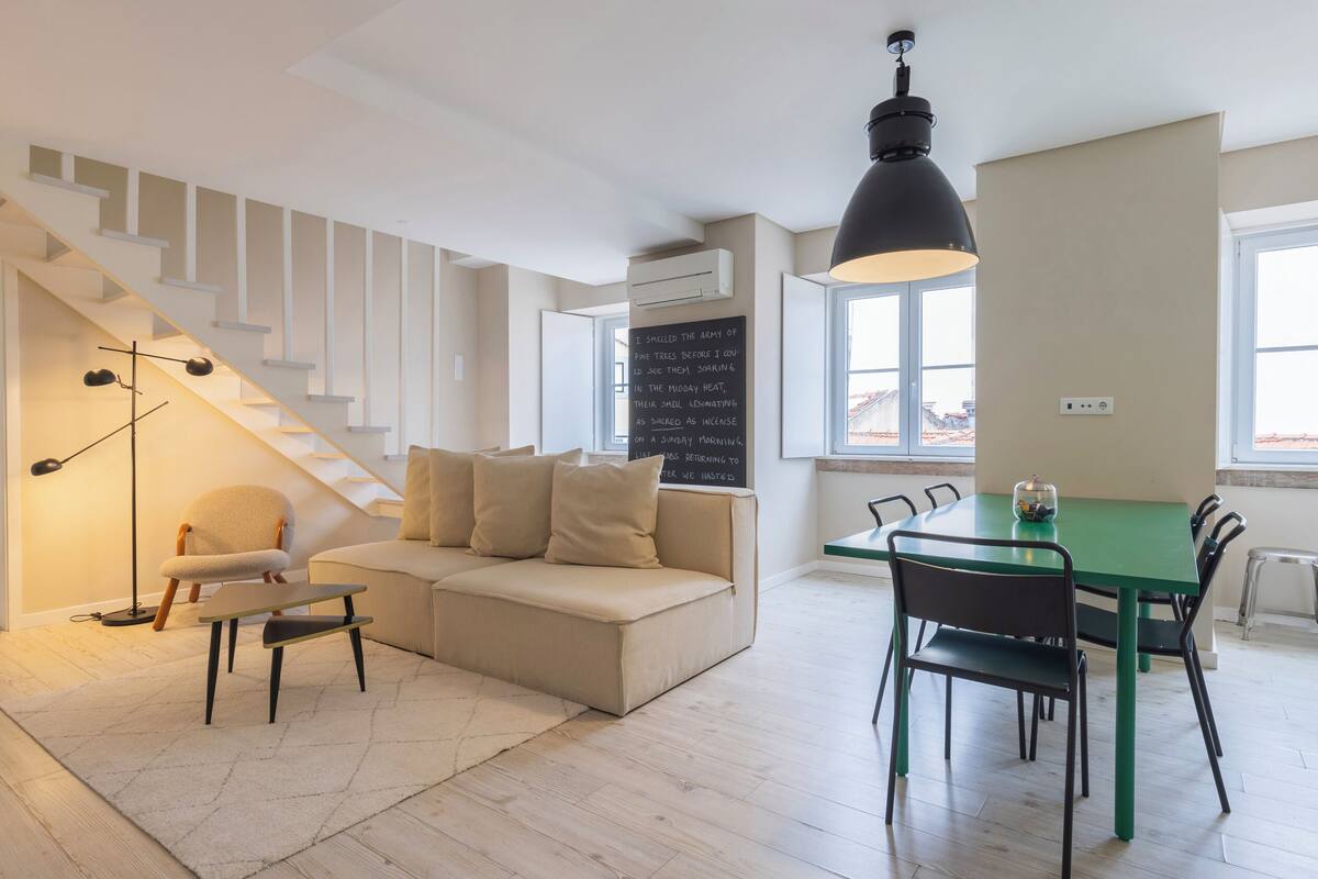 Una sala de estar moderna con una escalera, un sofá, una mesa de comedor con sillas y un ventanal grande.