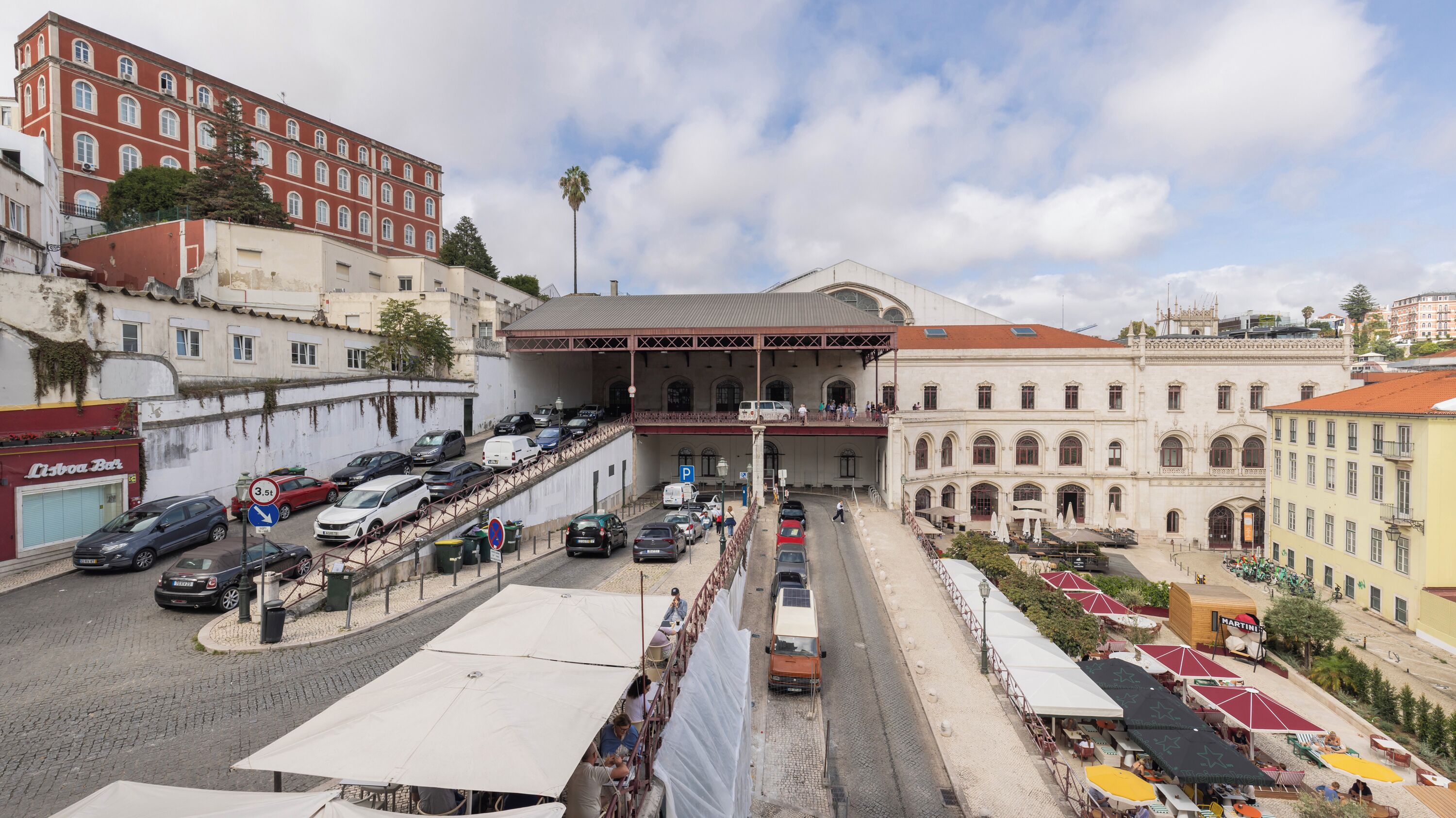 Vista de la calle con un edificio destacado, una galería techada y una plaza con mesas al aire libre.