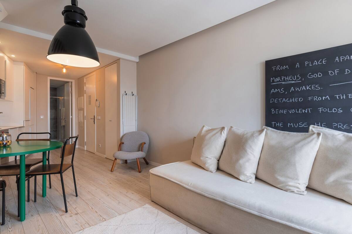 Una sala de estar moderna con una mesa de comedor verde, un sillón gris y un sofá beige. Un pizarrón con texto está montado en la pared.