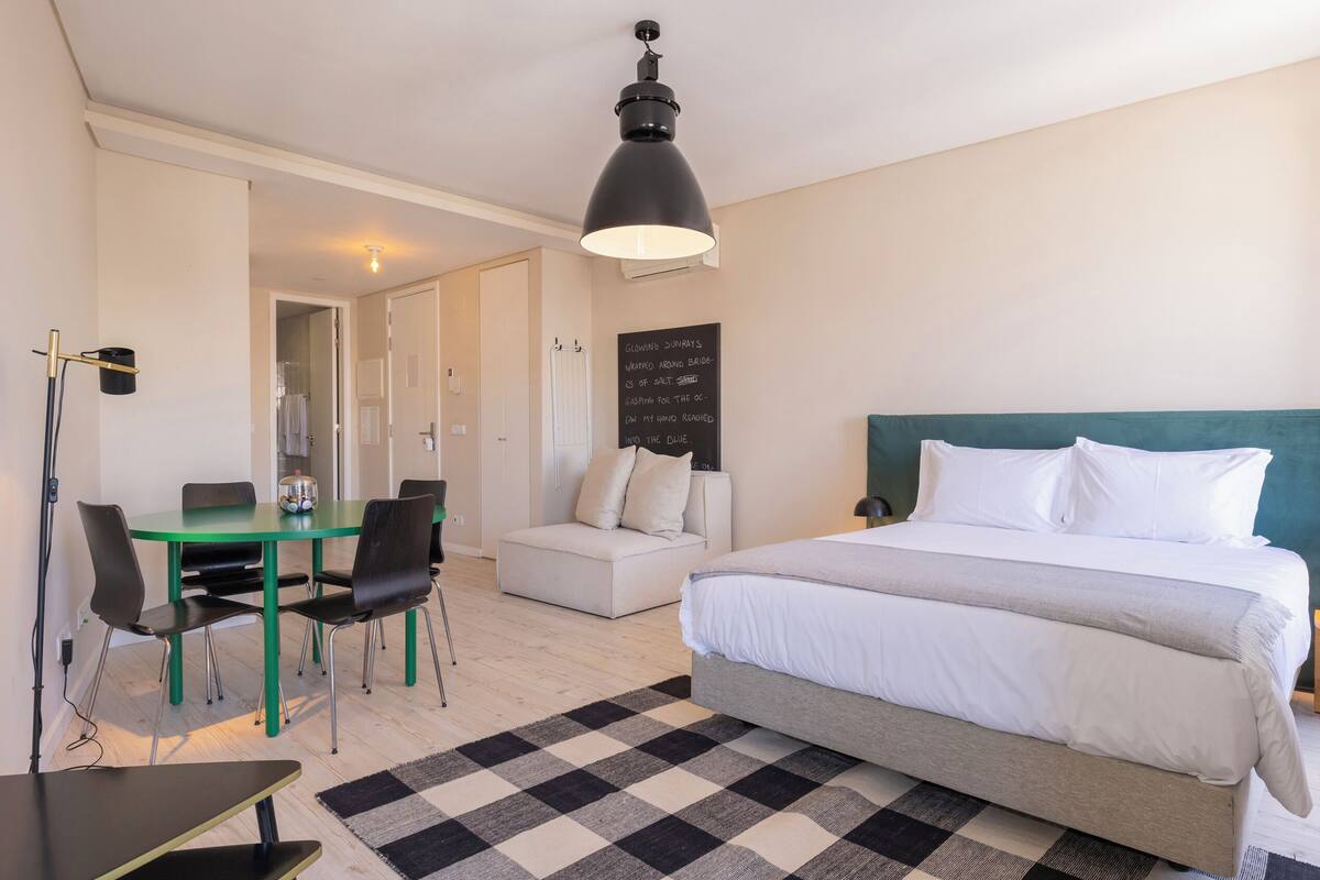 Habitación de hotel con una cama, un escritorio, sillas, una lámpara negra y una alfombra a cuadros.