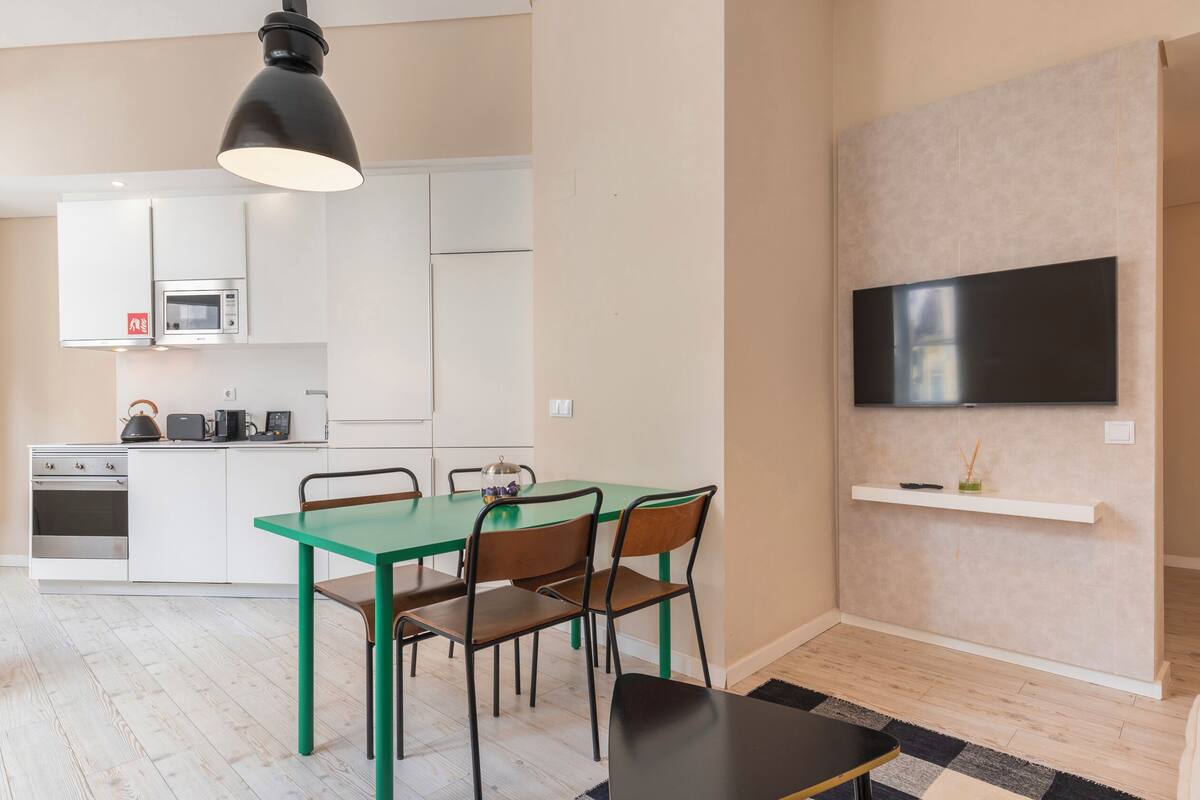 Una cocina moderna con armarios blancos, un microondas y una tetera. Hay una mesa de comedor con sillas y un televisor montado en la pared.