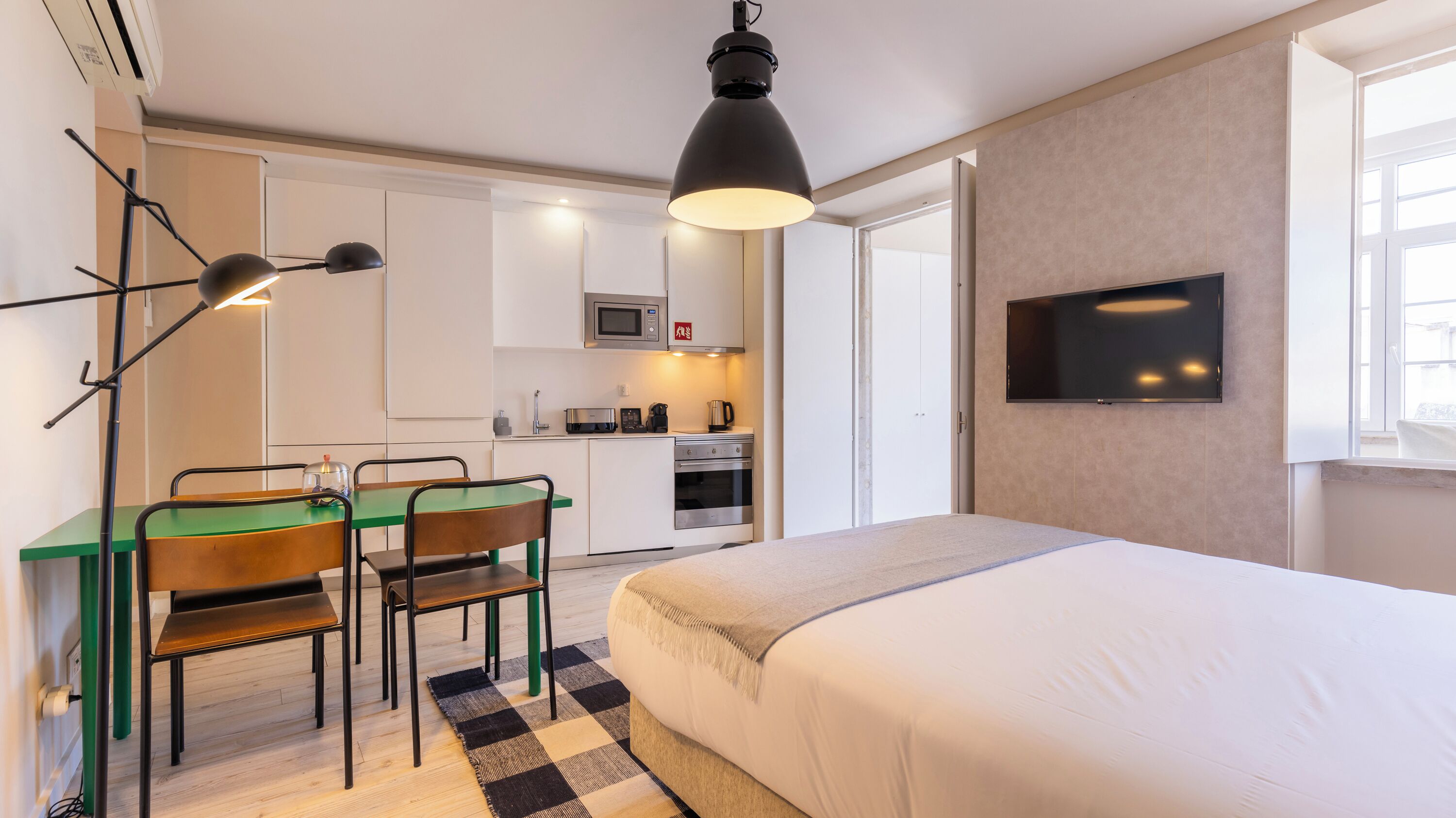 Une chambre d’hôtel moderne équipée d’un lit, d’un bureau avec des chaises, d’une kitchenette et d’un téléviseur fixé au mur.