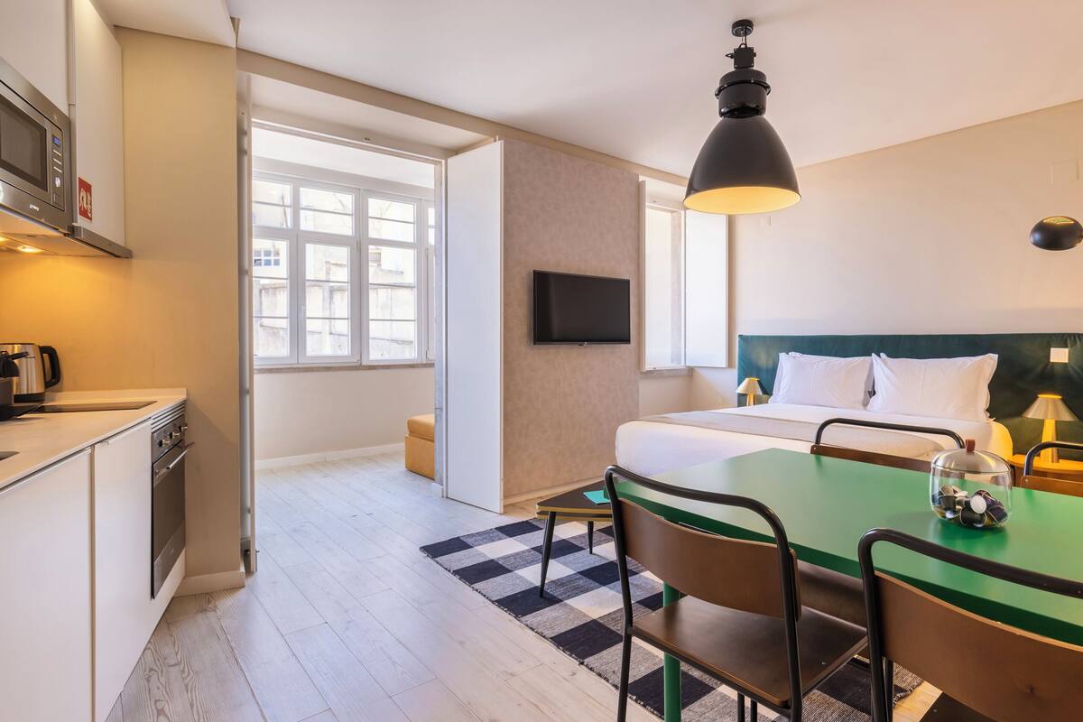 Una habitación de hotel moderna con kitchenette, una cama con ropa blanca, un comedor verde y un ventanal grande.