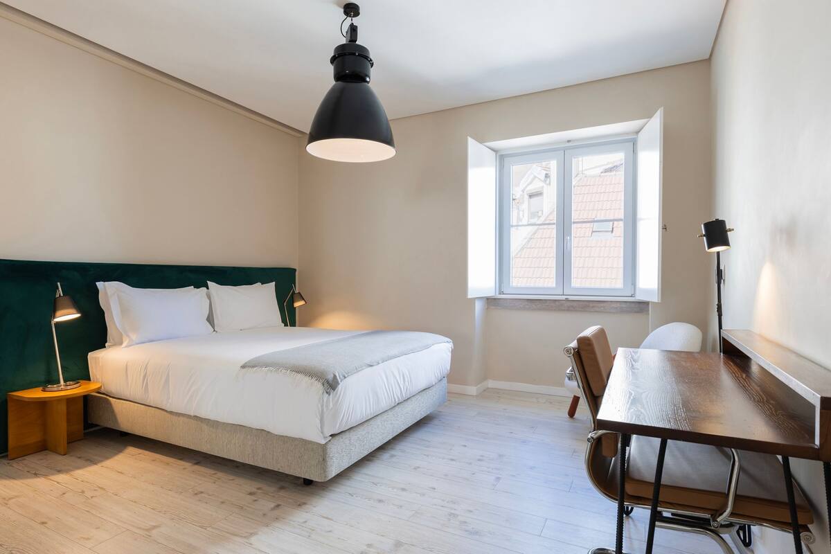 Un dormitorio con cama, mesita de noche, lámpara, silla, escritorio y ventana.