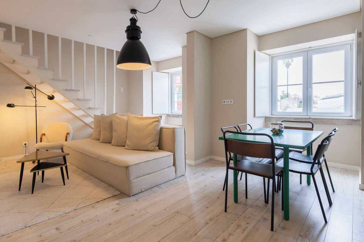 Una sala de estar moderna con una escalera, un sofá, una mesa de comedor con sillas y un amplio ventanal.
