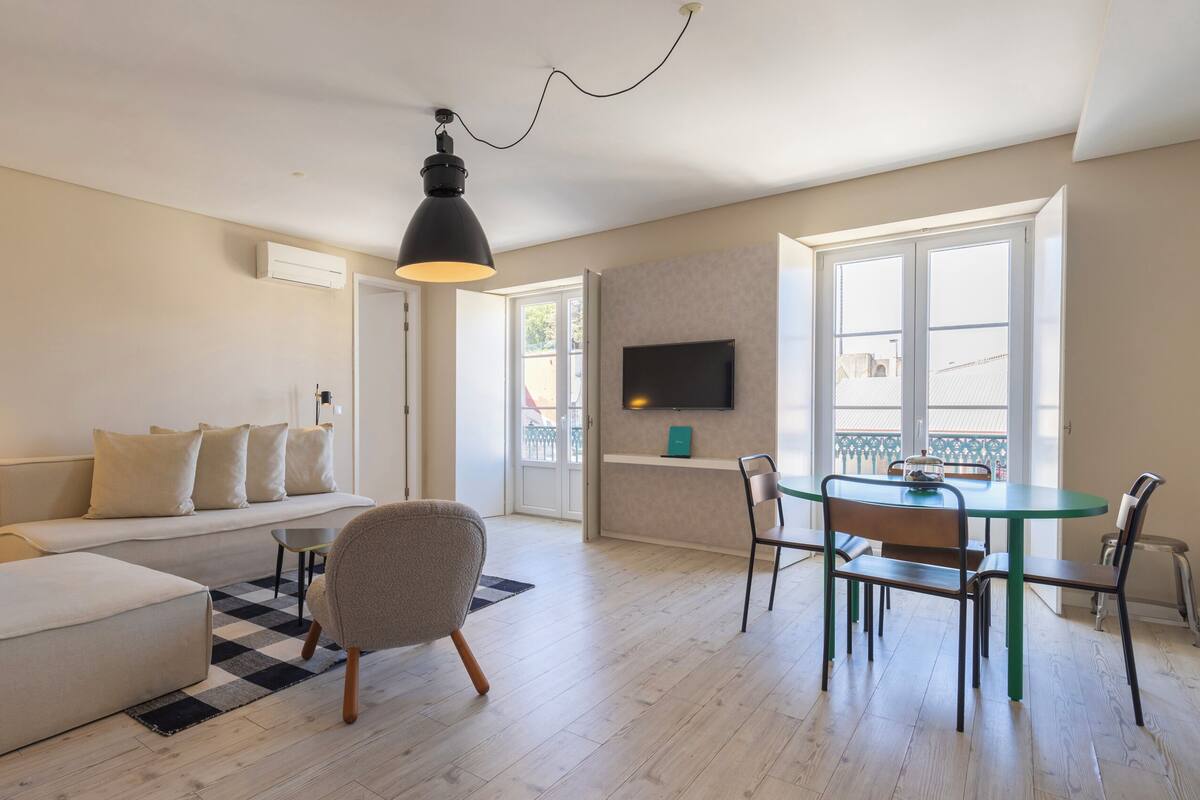 Una sala de estar moderna con sofá, sillón, mesa de comedor y sillas. Hay un televisor montado en la pared y un ventanal que proporciona luz natural.
