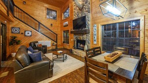 Un acogedor interior de cabaña de madera con chimenea, comedor, sofá de cuero y escalera.