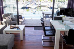 A modern living room with a view of a marina. It includes a sofa, a coffee table, a dining table, and a TV. - Hausboot in Boltenhagen (Boltenhagen)