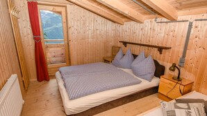 A wooden cabin bedroom with a bed, pillows, a nightstand, a window with a red curtain, and a view of mountains.