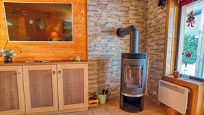 A room with a stone wall, wooden cabinets, a large TV, a wood-burning stove, and a window with a view. - Doppelhaushalfte in Hasselfelde (Oberharz am Brocken)