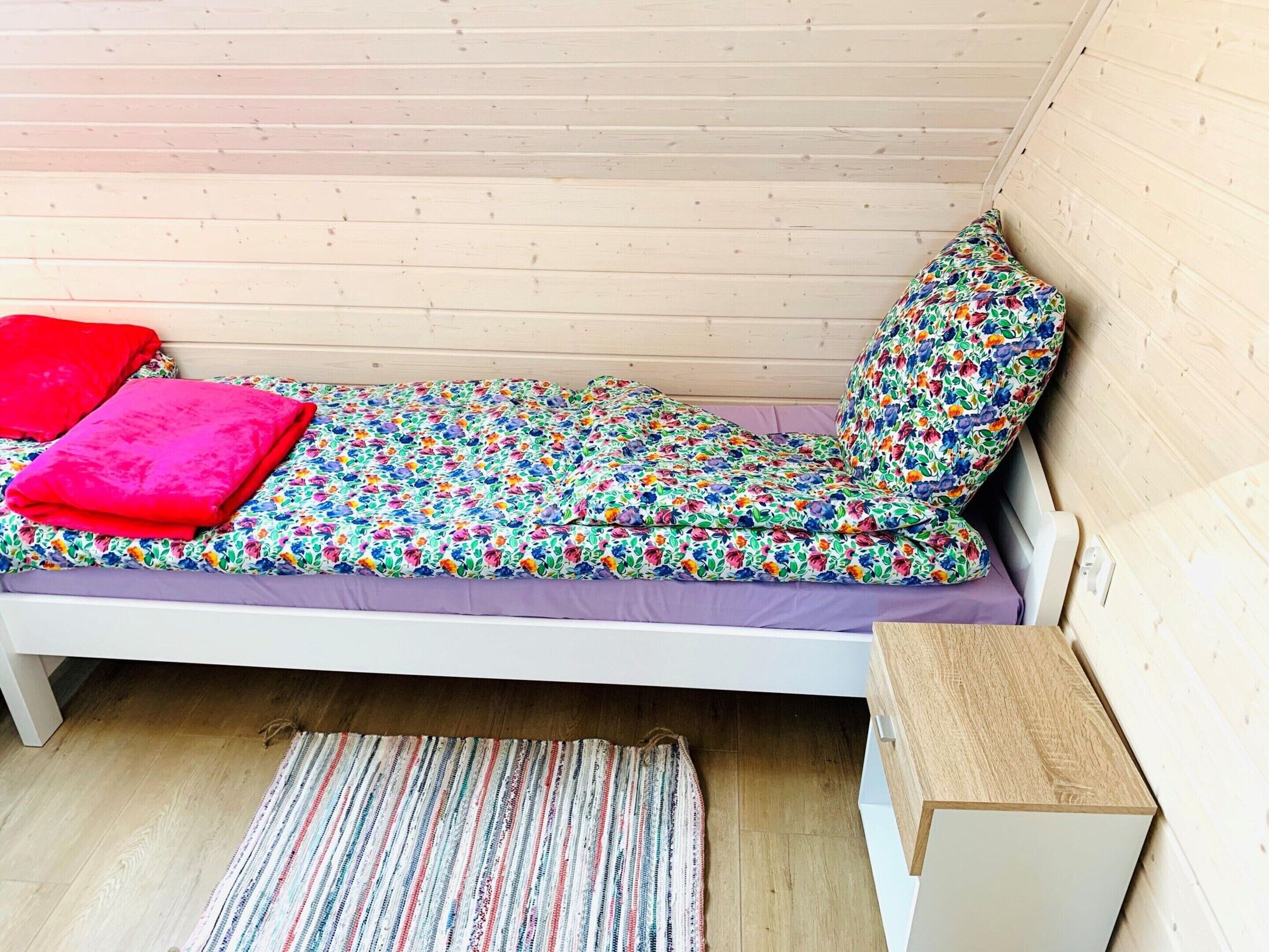 A small, single-bed room with a wooden sloped ceiling, a colorful floral bedspread, and a striped rug on the floor.