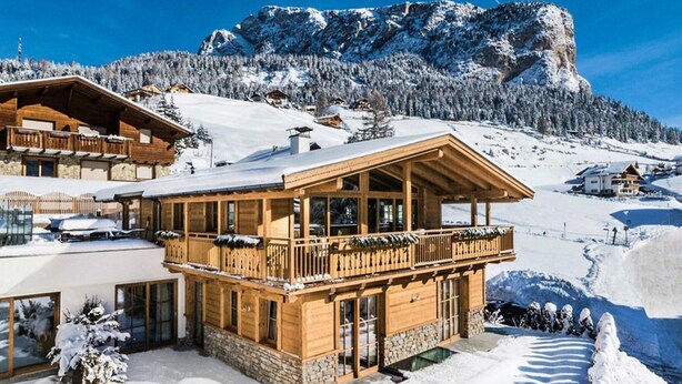 En træchalet med balkon, omgivet af sneklædte bjerge og klare himmel.
