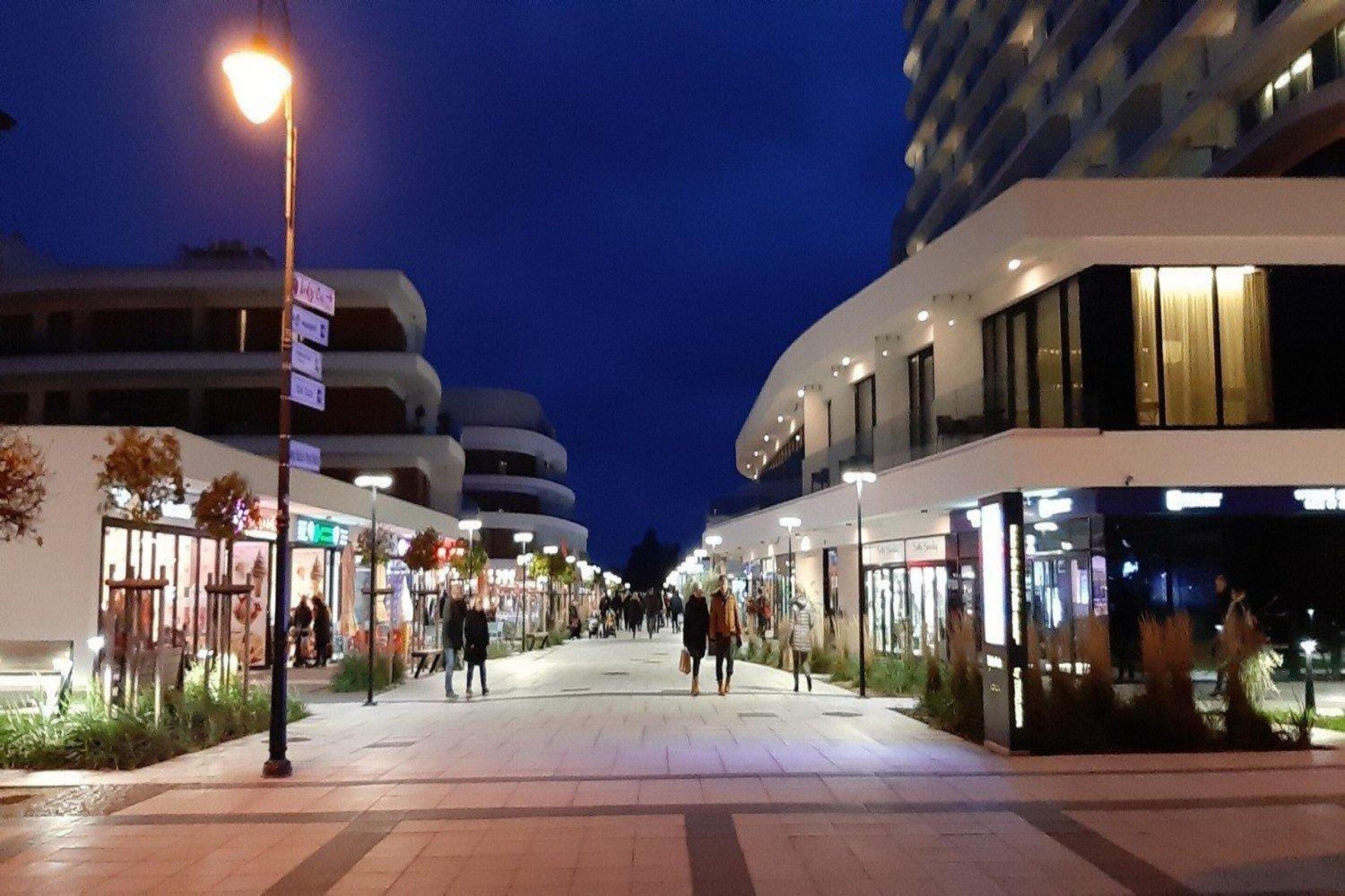 Calle urbana nocturna con edificios modernos, una acera para peatones y escaparates iluminados.