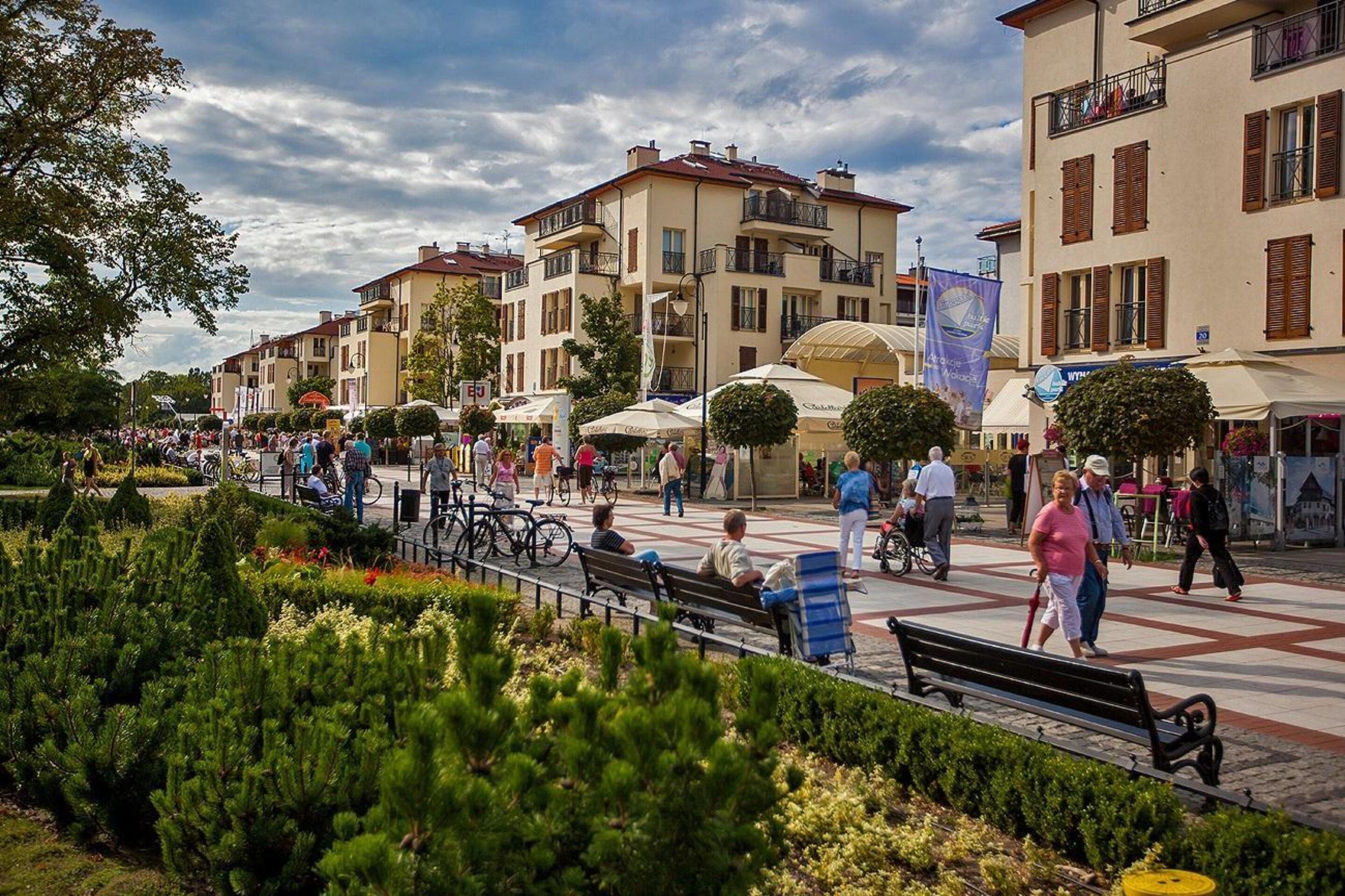 A public square with benches, people walking, and buildings with balconies.