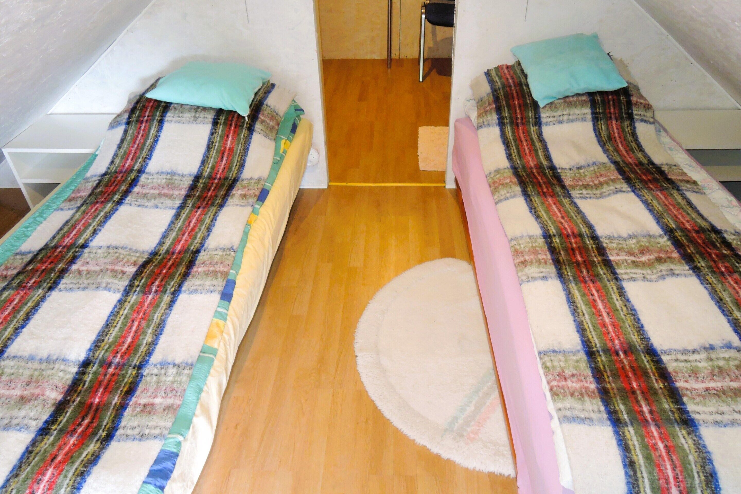 A room with two beds, each with a plaid blanket and a pillow. There is a wooden floor and a small rug in the center.
