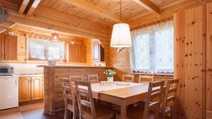A wooden cabin interior with a dining table, chairs, a kitchen area, and a large window with lace curtains. - Holiday Cottage in Pobierowo Near Baltic Beach (Rewal)