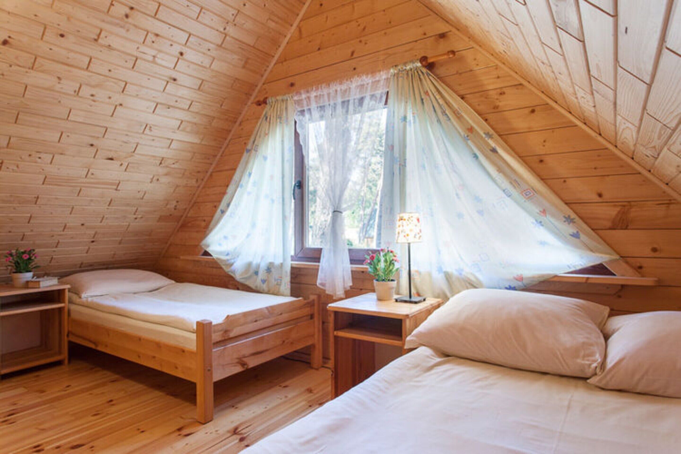 A cozy attic bedroom with two wooden beds, a small bedside table with a lamp, and a window with sheer curtains.
