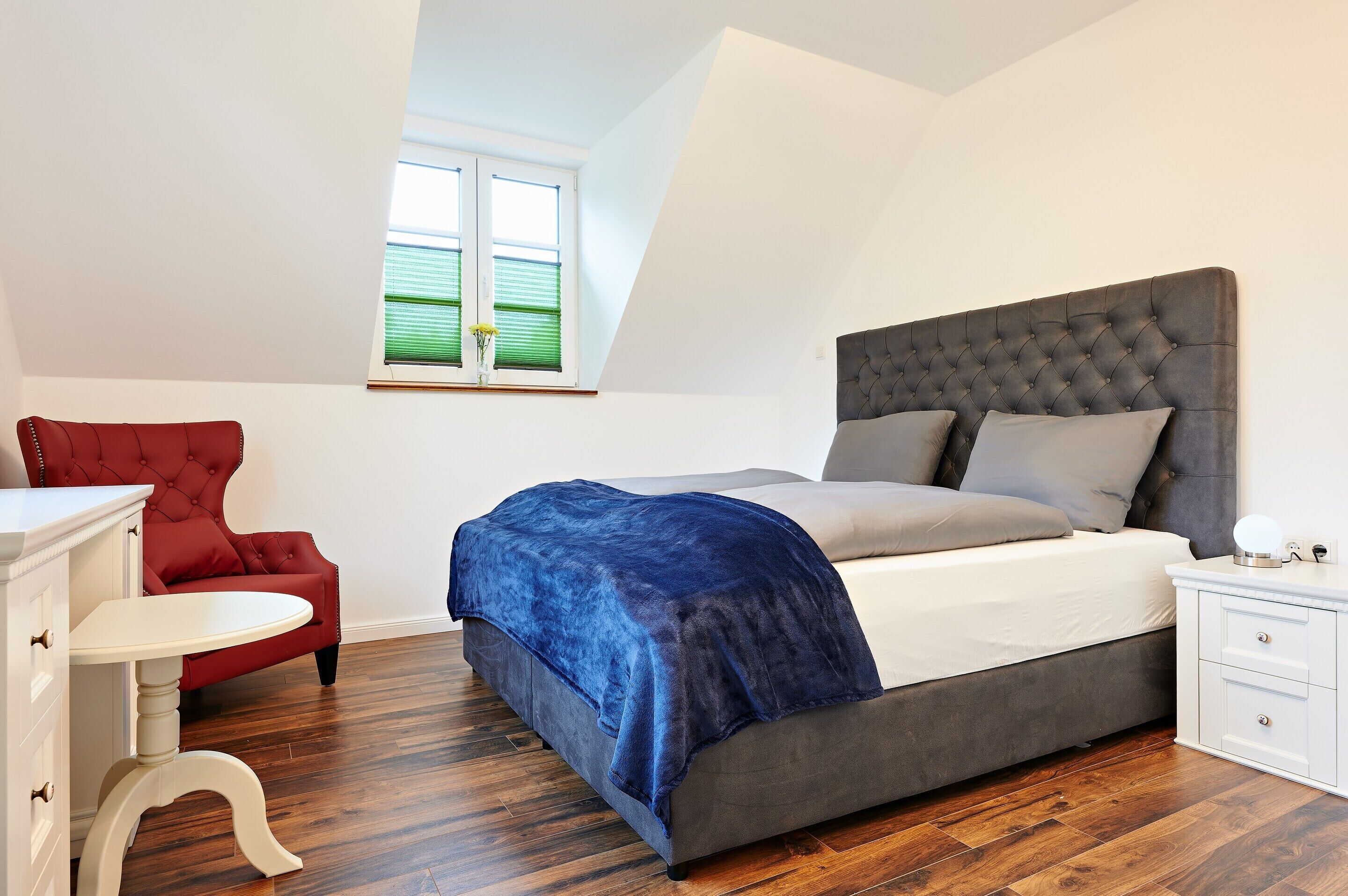 A bedroom with a bed, a red chair, a white nightstand, and a window with blinds.