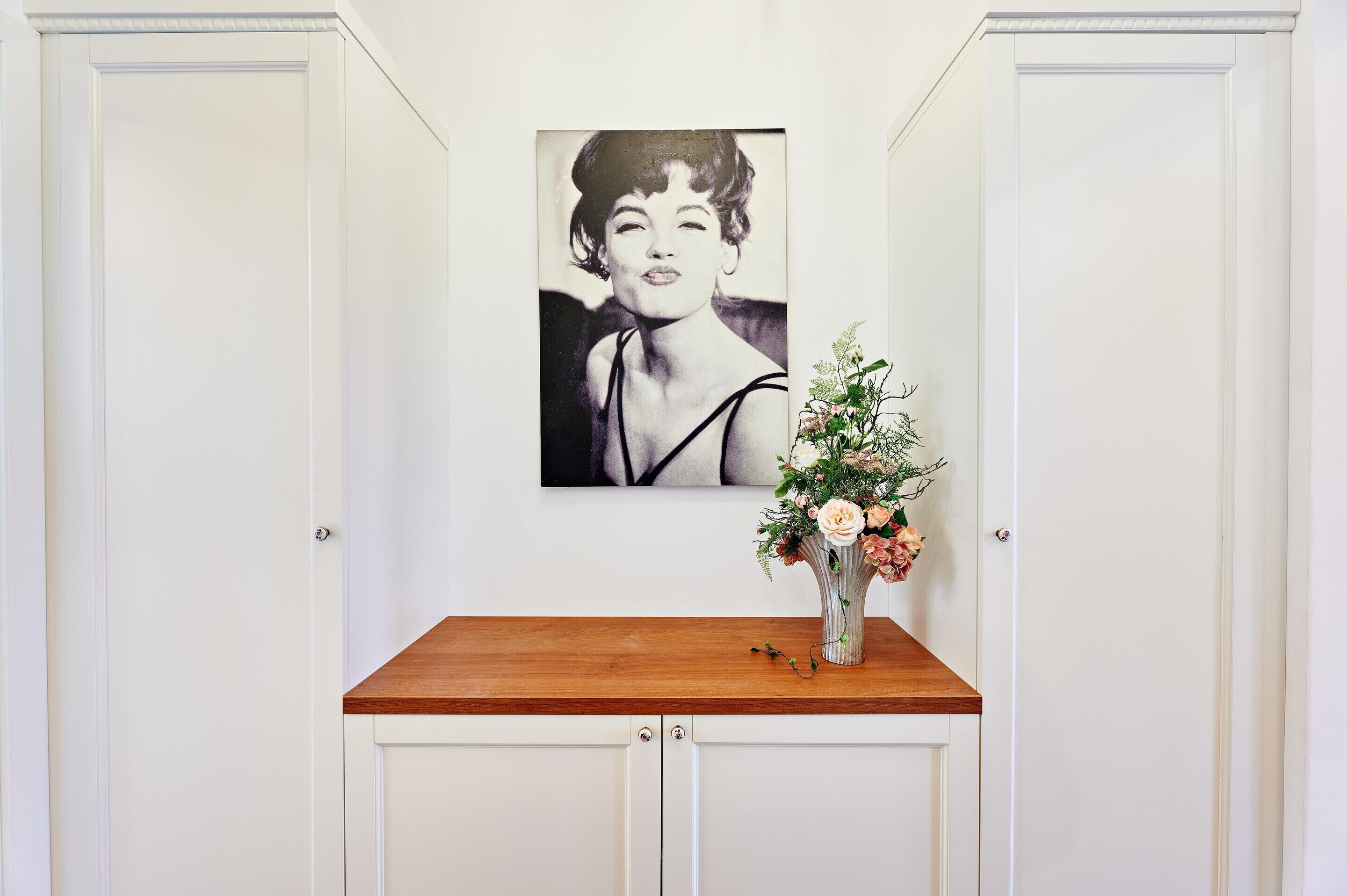 A white room with a wooden table, a black and white portrait, and a vase with flowers.