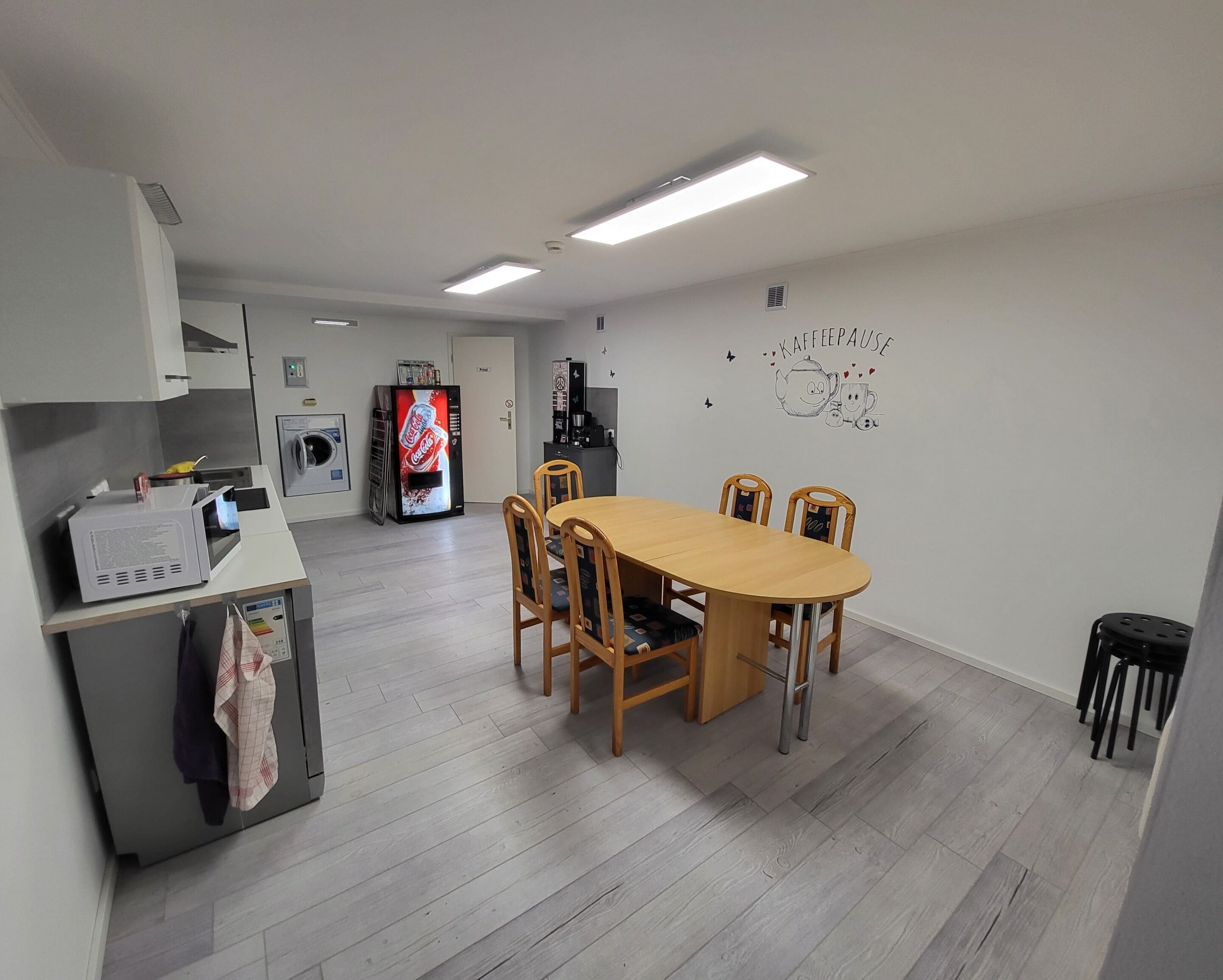 Een compacte keuken met een magnetron, een koelkast en een Coca-Cola koelkast. Er is een houten eettafel met stoelen en een wandgemonteerde koffiemachine.