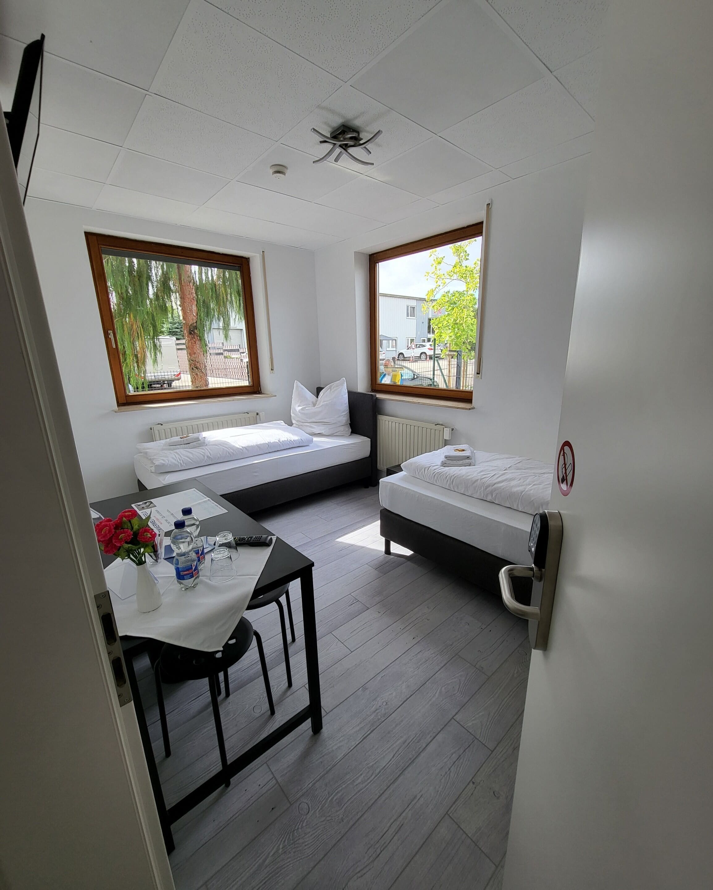 A small, clean room with two beds, a table with a vase of flowers, and a view of the street through large windows.