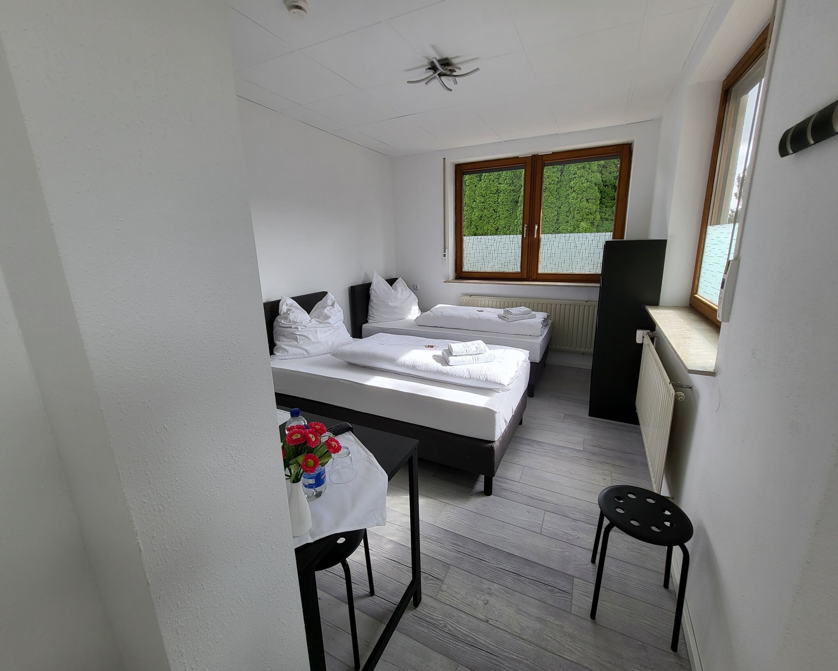 Ein kleines Hotelzimmer mit zwei Betten, einem Tisch mit Blumenvase, einem Sessel, einem Schreibtisch und einem Fenster mit Blick auf Grünflächen.