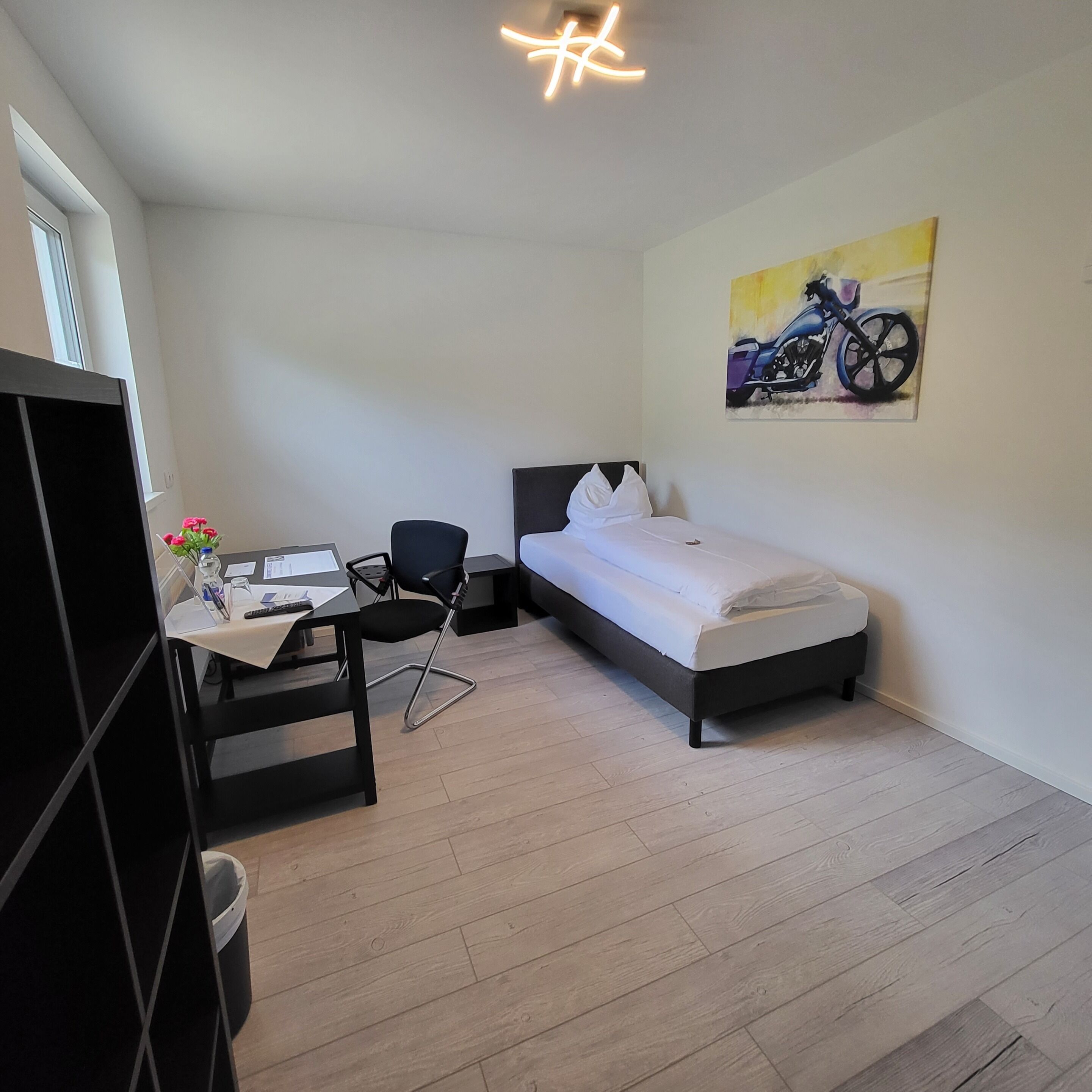 A small, modern room with a bed, a desk, and a chair. There is a painting on the wall and a window providing natural light.