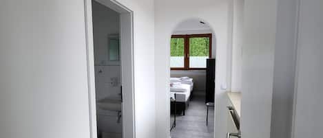 A narrow hallway with a view into a bedroom with a bed and a window.