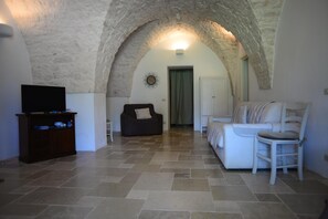 A living room with a stone archway, a television on a wooden cabinet, a sofa, and a white armchair. - Villa in Puglia Near Ostuni Marina (Ostuni)