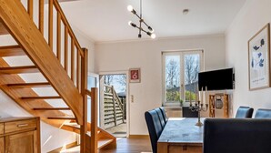 A dining area with a wooden staircase, a dining table with chairs, a TV mounted on the wall, and a window with a view of the outside.