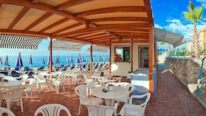A covered outdoor seating area with white plastic chairs and tables, overlooking the sea with blue umbrellas.